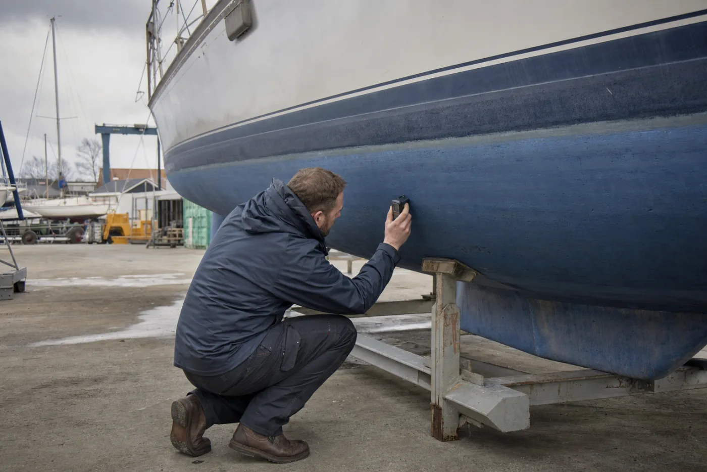 A surveyor examining a yacht hull ashore in grey weather